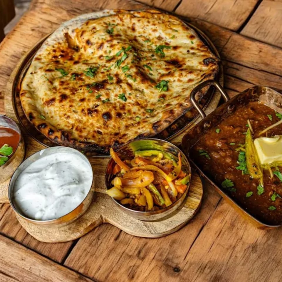 Indian meal with naan bread, dark curry, raita, and sautéed vegetables on a wooden table.