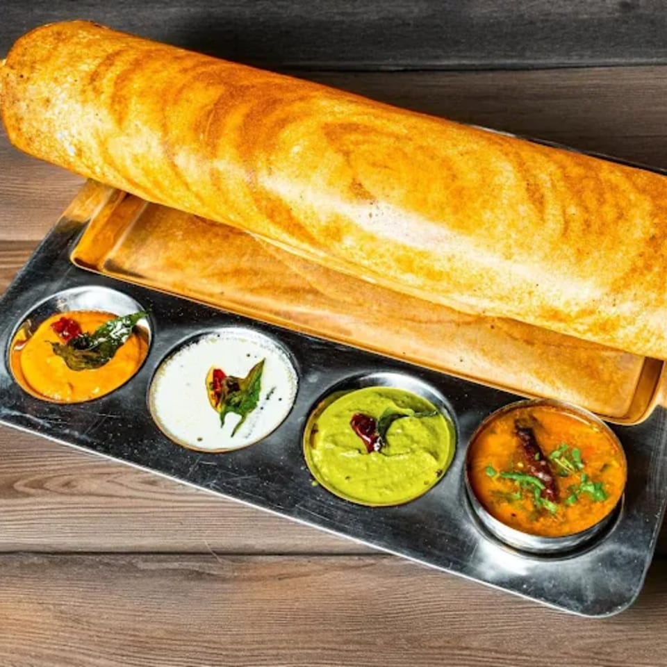 A golden-brown dosa is served on a metal tray with four small bowls of colorful Indian chutneys.